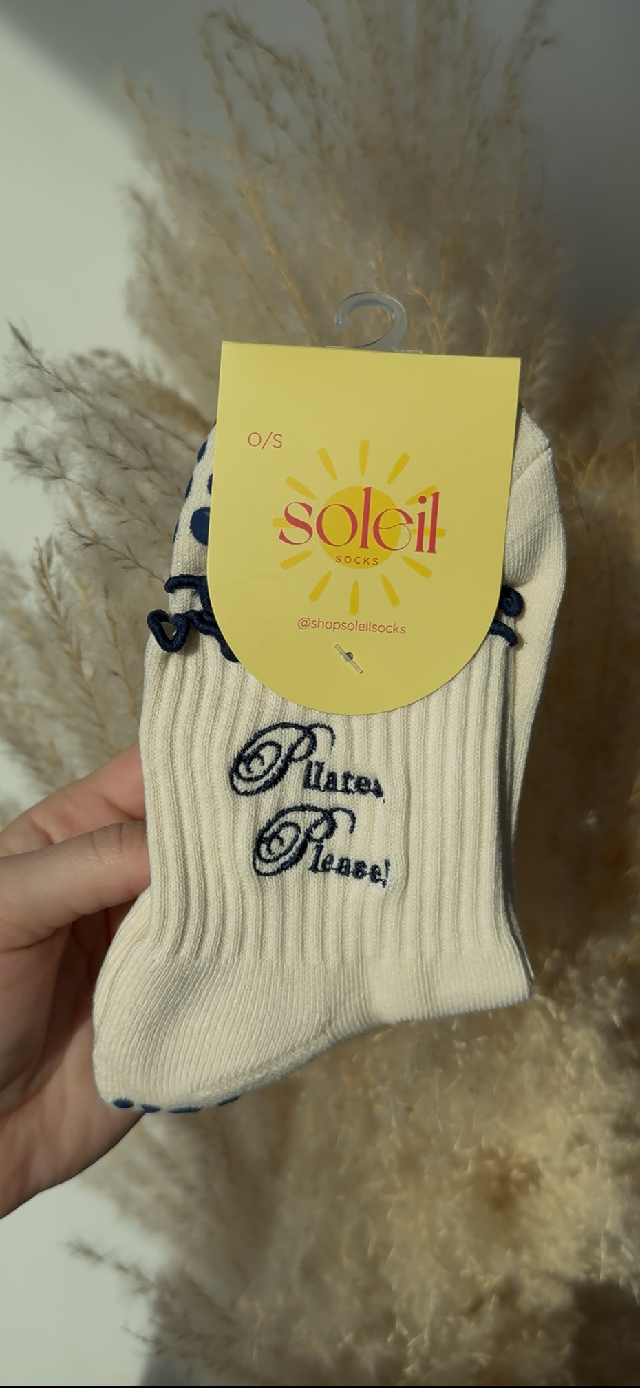 cream white sock with navy blue text "Pilates, please!"held by a hand against a textured wall background