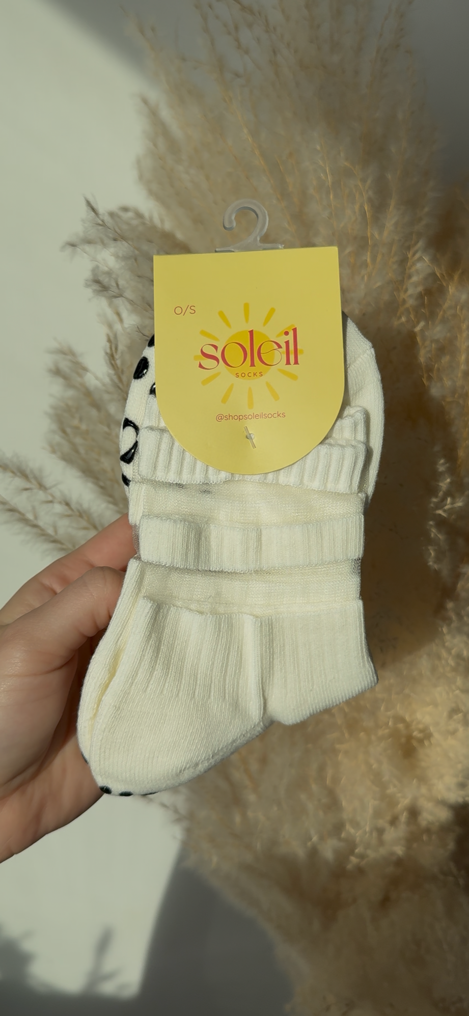 White grippy socks held by a hand with a 'soleil' tag against a decorative background.