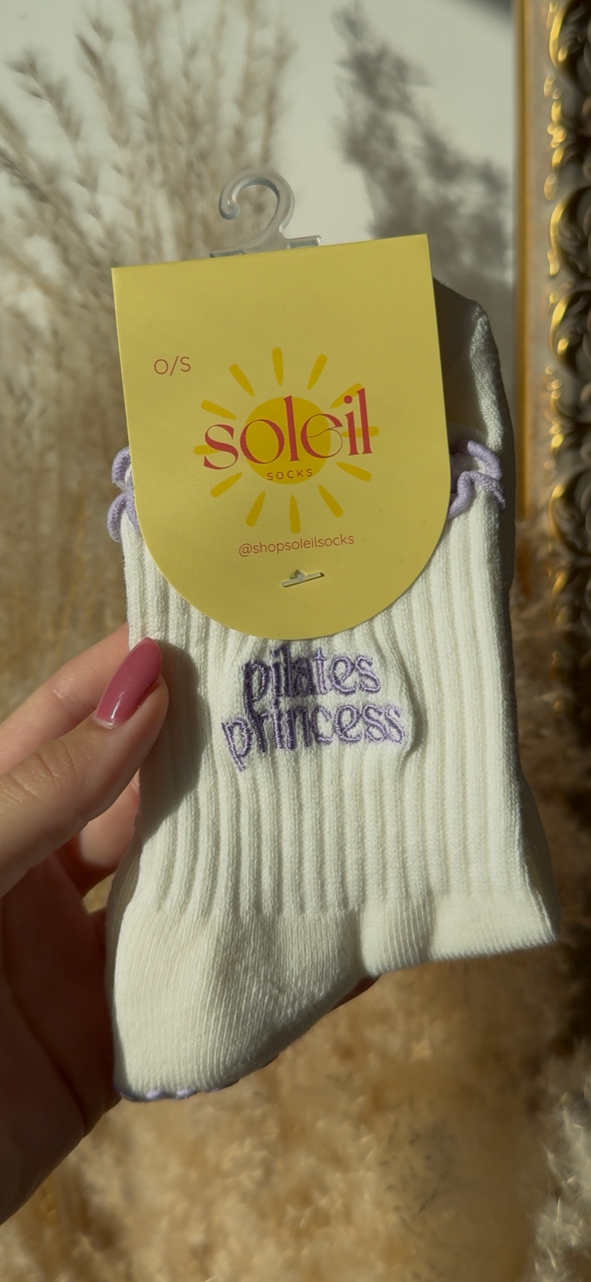 White socks with a purple text "pilates princess" yellow tag labeled 'soleil' held by a hand against a decorative wall.